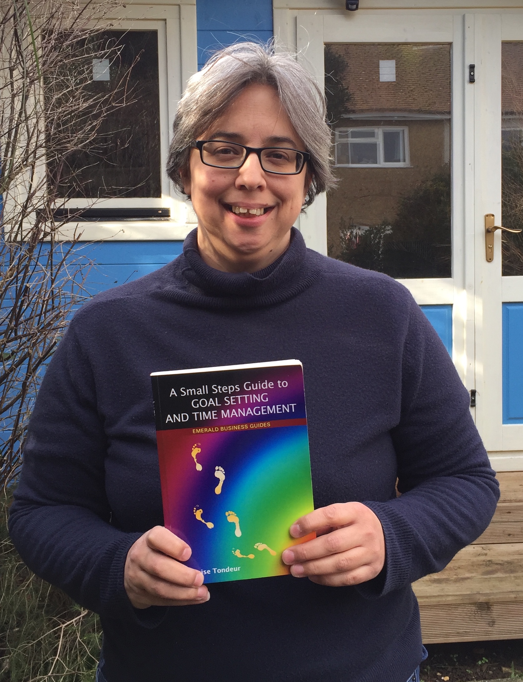 Photo of Lou outside her writing shed with Small Steps Guide to Goal Setting and Time Management